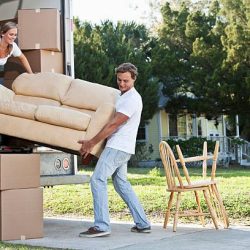 istockphoto-184100344-612×612 A young couple lifts a tan couch to move it in or out of the back of a moving truck. A young woman with blond hair pulled away from her face is standing inside the truck holding the left end of the couch. The woman wears a white top and dark blue jeans. A young man with brown hair stands outside the truck holding the right end of the couch. The man is wearing a white shirt and light blue jeans. Moving boxes and sit inside and outside the truck. Two chairs sit on a sidewalk outside the truck. A yellow house can be seen in the background.