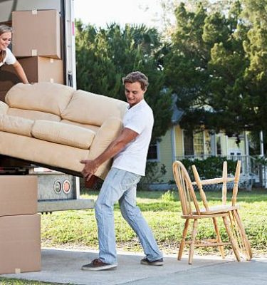 istockphoto-184100344-612×612 A young couple lifts a tan couch to move it in or out of the back of a moving truck. A young woman with blond hair pulled away from her face is standing inside the truck holding the left end of the couch. The woman wears a white top and dark blue jeans. A young man with brown hair stands outside the truck holding the right end of the couch. The man is wearing a white shirt and light blue jeans. Moving boxes and sit inside and outside the truck. Two chairs sit on a sidewalk outside the truck. A yellow house can be seen in the background.