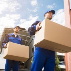 Close-up Of Two Young Delivery Men Carrying Cardboard Box In Front Of Truck Close-up Of Two Young Delivery Men Carrying Cardboard Box In Front Of Truck