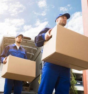 Close-up Of Two Young Delivery Men Carrying Cardboard Box In Front Of Truck